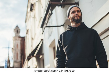 City Portrait Of Handsome Hipster Man With Beard Wearing Black Blank Hoodie Or Hoody And Hat With Space For Your Logo Or Design. Mockup For Print