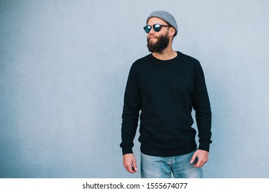 City Portrait Of Handsome Hipster Man With Beard Wearing Black Blank Hoody Or Sweatshirt And Hat With Space For Your Logo Or Design. Mockup For Print