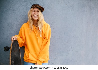 City portrait of positive young female wearing orange hoody and baseball cap holding skateboard. Grey blue wall on background  - Powered by Shutterstock