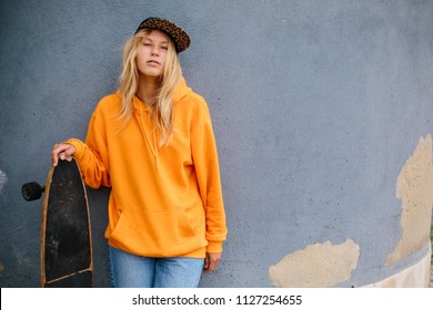 City portrait of positive young female wearing orange hoody and baseball cap holding skateboard. Grey blue wall on background 