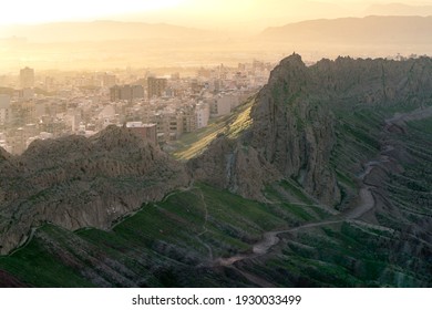 City of Qom, Iran, in a dramatic sunset viewed from the hill nearby. Steep rock cliffs in the front. Desert rocks, dramatic terrain.