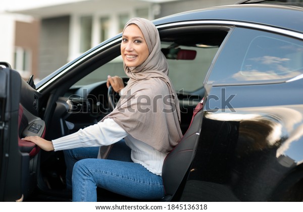 City Ride Concept. Portrait Of Happy Smiling\
Muslim Lady In Hijab Getting In Or Out Of Car, Holding Open Door.\
Arabian Woman In Headscarf Driving Modern Vehicle, Looking Back And\
Posing At Camera