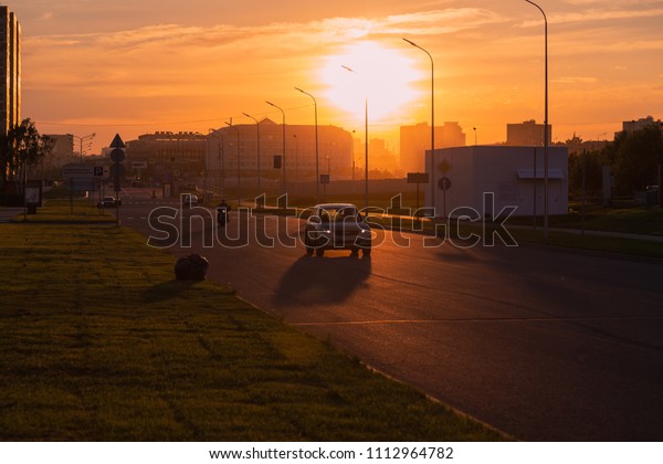City road at sunset on which the car and
motorcycle rides. Warm spring evening. Green lawns lit by orange
rays of the sun. On the roadside is a black polyethylene bag with a
beveled grass lawn mower.