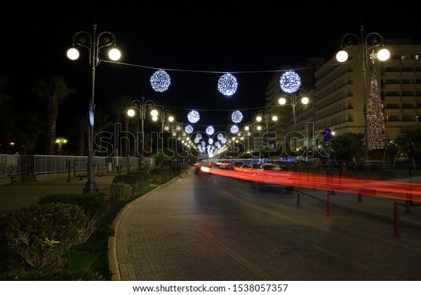 City street at night,\
Christmas time, high decorative Christmas tree, illuminated\
decorations hang on cables  in air, buildings, trails of red lights\
left by car in motion