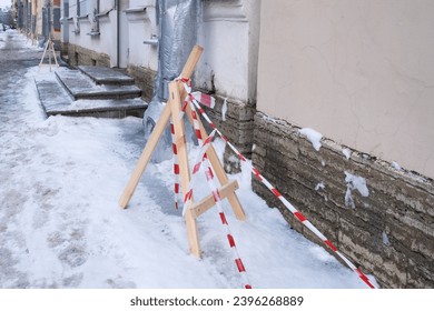City streets after snowfall. Snow-covered sidewalks are fenced off with warning tape on wooden pyramids. Melting snow on water pipes freezes into ice and icicles that are dangerous for pedestrians