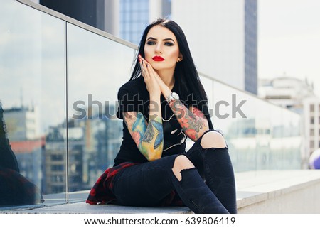 City style. Portrait of attractive young tattooed hipster girl touching her face while sitting against urban background.