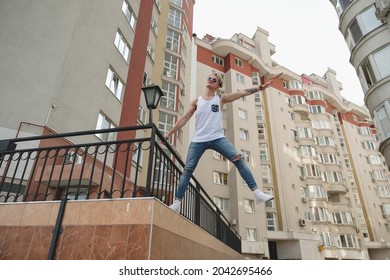 City Youth Concept. Boy Dreaming Of Freedom Hanging In Air One Arm And One Leg. Young Man In The City On The Buildings Background.