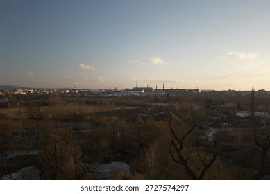 A cityscape appears distant. Fields stretch toward buildings. Bare trees populate the foreground. A cloudy sky dominates. Structures rise in the background - Plzen, Czech - Jan,18,2026