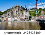 Cityscape with citadel on top of rocky mountain and collegiate church of Our Lady of Dinant, bridge with waving flags, buildings facing river Meuse, sunny summer day in Namur, Wallonia, Belgium