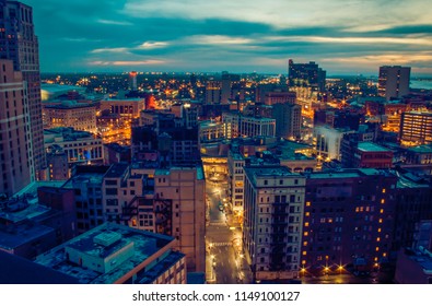 Cityscape of Detroit Michigan glowing at sunrise. Historical architecture, business buildings, apartments, skyscrapers and other structures in foreground. Cloudy blue gray sky with hints of orange.