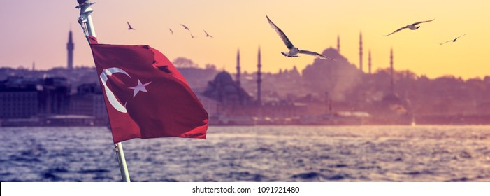 Cityscape of Istanbul with silhouettes of ancient mosques and minarets at sunset. Panoramic view on the old city and flying seagulls on blur background with turkish flag and ship in front.