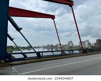 A cityscape viewed through the structural elements of an industrial bridge, highlighting red and blue beams against an overcast sky, with reflections on the river below. - Powered by Shutterstock