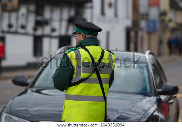 civil\
enforcement officer traffic warden issues ticket to black car at\
roadside with typical English town in\
background