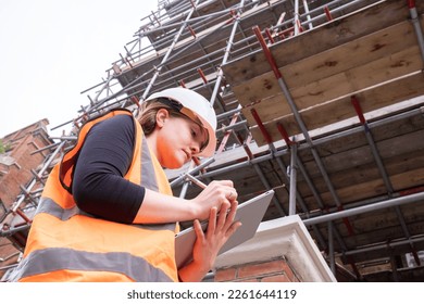 Civil engineer lady writing on a tablet with an electronic pen, hard hat and orange personal protective equipment, survey, touchscreen, technology and innovation in construction site, low angle view - Powered by Shutterstock