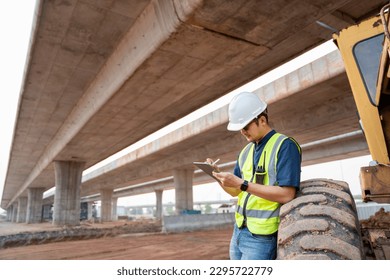 A civil engineer logging data with ipad standing beside industrial grader wheels at road construction site.