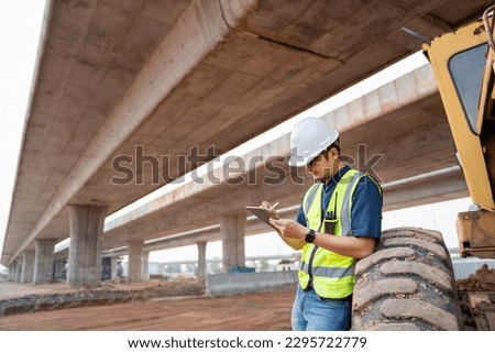 A civil engineer logging data with ipad standing beside industrial grader wheels at road construction site.