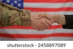 A civilian woman and a military man shake hands in front of an american flag, symbolizing collaboration.