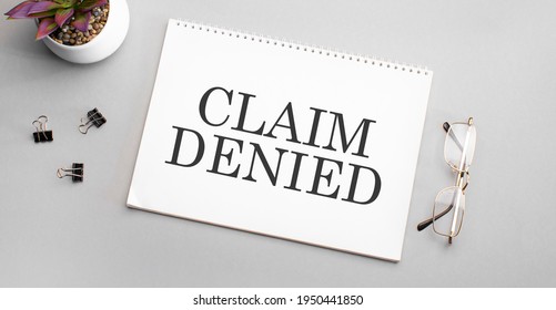 Claim Denied is written in a white notebook next to a pencil, black-framed glasses and a green plant. - Shutterstock ID 1950441850