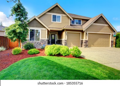 Clapboard Siding House  With Stone Trim. View Of  Entrance Porch, Beautiful Flower Bed With Green Lawn, Bushes And Fir Tree