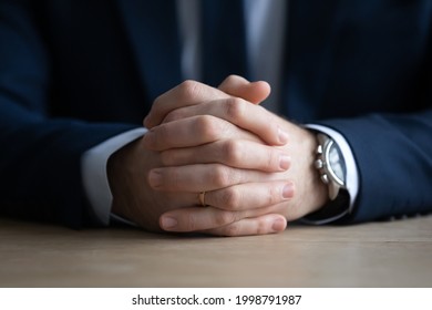 Clasped hands of businessman in formal suit sitting at work or meeting table. Attentive professional taking part in interview, negotiation, ready for dialogue, conversation. Cropped close up