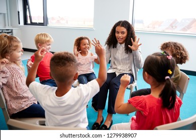 A class of infant school children sitting on chairs in a circle in the classroom, raising hands and learning to count with their female teacher, close up