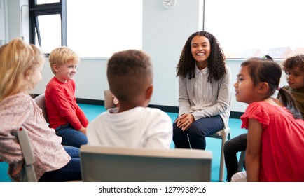 A Class Of Infant School Children Sitting On Chairs In A Circle In The Classroom Talking To Their Female Teacher