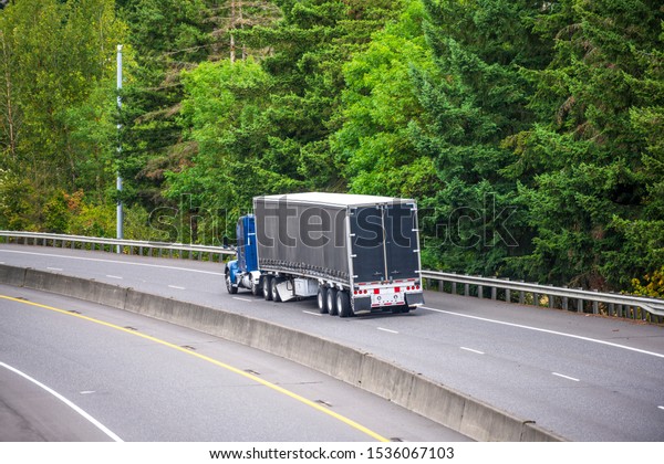 Classic\
American blue bonnet big rig semi truck with sleeping compartment\
transporting cargo in black covered semi trailer running on the\
divided highway with green trees on the\
side