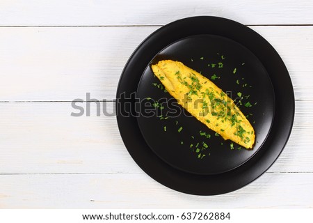 classic basic French three eggs fluffy omelette sprinkled with finely chopped parsley on black plate,  on white wooden table, view from above 