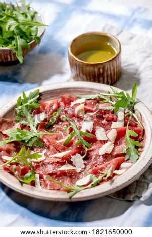 Classic beef carpaccio with cheese and arugula on ceramic plate, served with olive oil over white and blue tablecloth.
