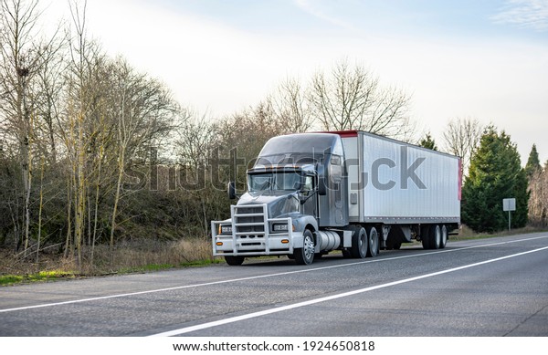 Classic big rig gray semi\
truck tractor with grille guard transporting commercial  cargo in\
dry van semi trailer standing on the highway road shoulder out of\
service