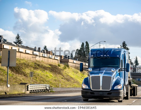 Classic big rig industrial blue semi truck tractor
with low roof cab and sleeping compartment transporting cargo on
step down semi trailer running on the wide highway with hill and
traffic road signs