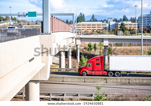 Classic big rig red semi truck tractor with\
sleeping compartment transporting commercial  cargo in dry van semi\
trailer moving on the highway intersection with overpass road and\
bridge with traffic