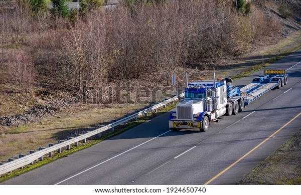 Classic big\
rig white semi truck tractor with low roof cab and sleeping\
compartment transporting empty step down semi trailer with oversize\
load signs moving on the one way highway\
road