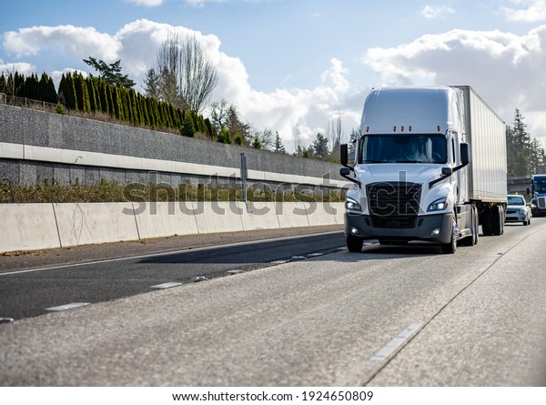 Classic big rig white semi truck tractor with\
sleeping compartment transporting commercial  cargo in dry van semi\
trailer moving on the highway road intersection with one way\
traffic direction