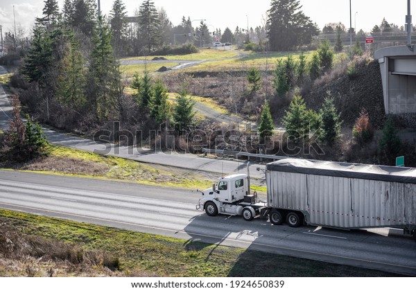 Classic big rig white semi truck tractor with day\
cab transporting commercial  cargo in covered bulk semi trailer\
moving on the highway road intersection with one way traffic\
direction and exit fork