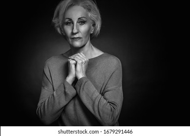 Classic black-and-white dramatic portrait of an elderly blonde woman in Studio on black background. hands folded on rude, the concept of supplication, uncertainty