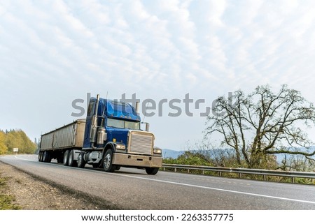 Classic blue big rig American bonnet powerful semi truck driving with covered bulk ribbed semi trailer transporting commercial cargo on the winding autumn road