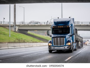 Classic Blue Long Haul Big Rig Bonnet Semi Truck Tractor Transporting Fixed With Slings Heavy Commercial Cargo On Flat Bed Semi Trailer Running On The Wet Highway Road At Rain Weather Condition