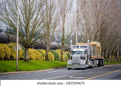 Classic bonnet commercial American big rig semi truck with flat bed semi trailer transporting containers with glass driving on the road with trees and bushes for timely delivery goods to warehouse 