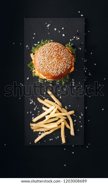 Classic burgers with becon, meat, cheese, onion,\
tomato and cheese and integral bun. On textured black board. Fried\
potatos. Flat lay.\
