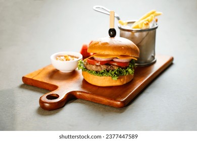 Classic Cajun Beef burger include cheese slice, tomato, onion and lettuce leaf served on wooden board with dip and french fries bucket isolated on grey background side view of appetizer fast food