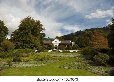 Classic Cape Dutch Farm House With Garden In The Foreground And Cloudy Blue Skies Overhead