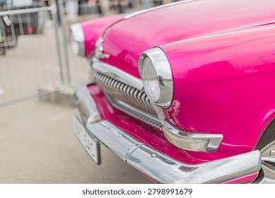 Classic car front section showcasing vibrant pink paint, chrome grille with vertical slats, circular headlights, polished bumper highlighting well-maintained condition against outdoor background.
