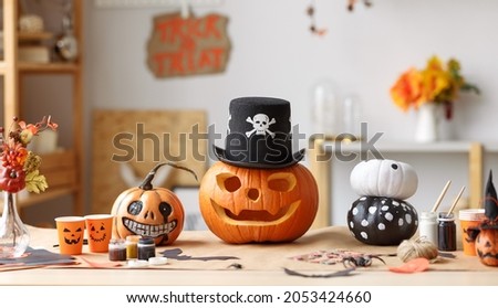 Classic carven spooky jack o lantern with big, gape-toothed grin in pirate hat standing on wooden table with different handmade halloween decorations and painted pumpkins, no people