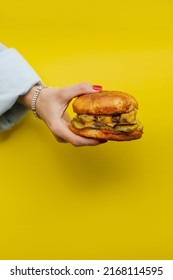 Classic Cheeseburger In A Woman’s Hand On A Bright Yellow Background. Juicy Appetizing Cheeseburger In Hand And Girls. Girl Holding A Burger Close Up