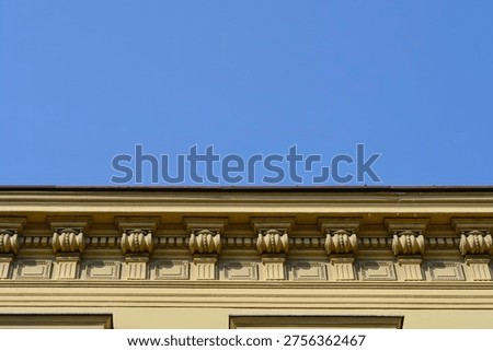 Classic decorative consoles and rectangular patterns adorn the upper cornice of a yellow historic building against a clear blue sky