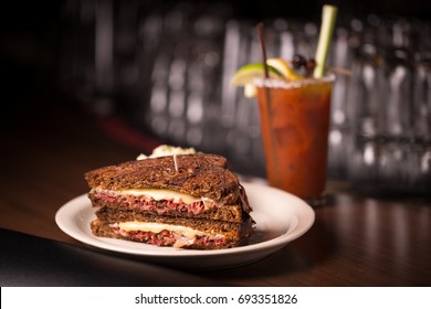 Classic Deli Or Diner Style Ruben Sandwich With Sliced Corned Beef Topped By Sauerkraut Coleslaw And Melted Swiss Cheese On Rye Bread With Hangover Bloody Mary Cocktail In Blurry Background