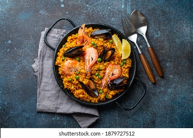 Classic dish of Spain, seafood paella in traditional pan on rustic blue concrete background top view. Spanish paella with shrimps, clamps, mussels, green peas and fresh lemon wedges from above 