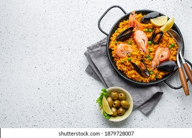 Classic dish of Spain, seafood paella in traditional pan on white wooden background top view. Spanish paella with shrimps, clamps, mussels, green peas and fresh lemon wedges from above, space for text