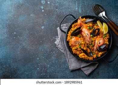 Classic dish of Spain, seafood paella in traditional pan on blue stone background top view. Spanish paella with shrimps, clamps, mussels, green peas and fresh lemon wedges from above, space for text